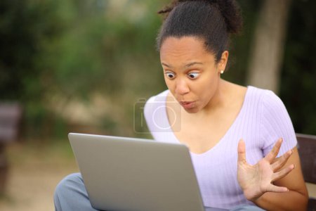 Téléchargez les photos : Surpris femme noire vérifier le contenu de l'ordinateur portable assis sur un banc dans un parc - en image libre de droit