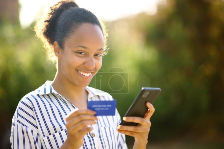 Photo for Black woman looking at you holding credit card and phone in a park - Royalty Free Image