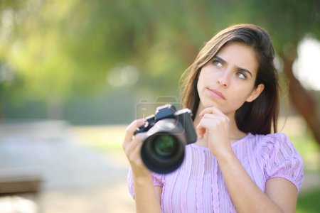 Téléchargez les photos : Photographe douteux pensant quoi photographier tenant la caméra dans un parc - en image libre de droit