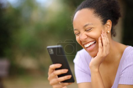 Téléchargez les photos : Femme noire drôle vérifiant téléphone riant fort dans un parc - en image libre de droit