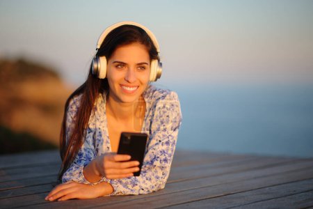 Foto de Mujer feliz con auriculares tumbados al atardecer escuchando música en el teléfono en la playa y contemplando - Imagen libre de derechos