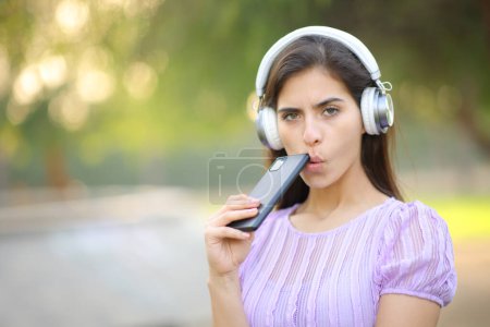 Téléchargez les photos : Femme suspecte vous regardant écouter audio numérique avec casque et smartphone dans un parc - en image libre de droit