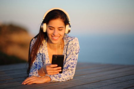 Téléchargez les photos : Portrait d'une femme heureuse au coucher du soleil écoutant un livre audio couché sur une terrasse en bois sur la plage - en image libre de droit