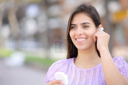 Téléchargez les photos : Femme heureuse mettant oreillette debout dans la rue regardant loin - en image libre de droit