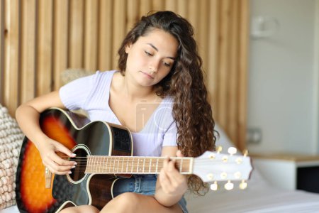 Téléchargez les photos : Femme jouant de la guitare assise sur le lit dans une pièce - en image libre de droit