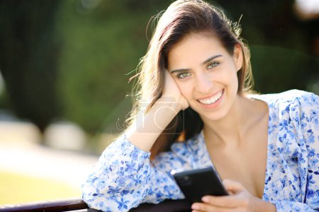 Foto de Hermosa mujer feliz posando mirando a la cámara sosteniendo el teléfono en un parque - Imagen libre de derechos