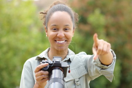 Photo for Front view portrait of a black photographer pointing at camera holding mirrorless camera in a park - Royalty Free Image