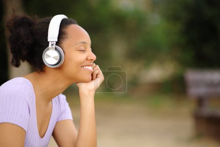 Téléchargez les photos : Heureuse femme noire relaxante écoute audio avec casque et les yeux fermés dans un parc - en image libre de droit