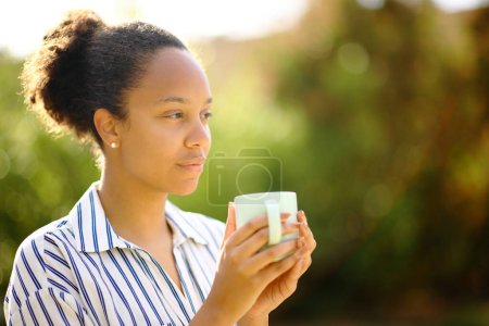 Foto de Relajada mujer negra sosteniendo taza de café mira hacia otro lado en un parque - Imagen libre de derechos