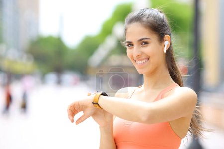 Photo for Runner wearing earbud and smartwatch selecting music looking at you in the street - Royalty Free Image