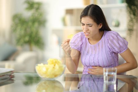 Photo for Woman suffering stomach ache eating chips at home - Royalty Free Image