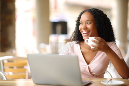 Téléchargez les photos : Heureuse femme noire avec ordinateur portable buvant du café et riant regardant loin dans une terrasse du restaurant - en image libre de droit