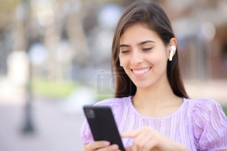 Téléchargez les photos : Femme heureuse avec écouteur et téléphone marchant dans la rue - en image libre de droit