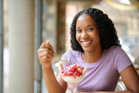 Foto de Feliz mujer negra comiendo postre en una terraza del restaurante mirando a la cámara - Imagen libre de derechos