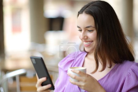 Téléchargez les photos : Joyeuse femme vérifiant téléphone portable et souriant dans une terrasse de café - en image libre de droit