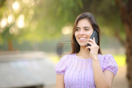 Téléchargez les photos : Femme heureuse appelant au téléphone marchant seule dans un parc verdoyant - en image libre de droit