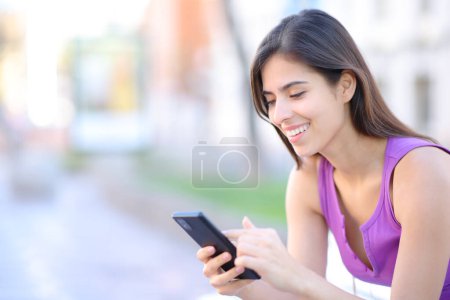 Téléchargez les photos : Femme heureuse utilisant un téléphone portable assis sur un banc dans la rue - en image libre de droit