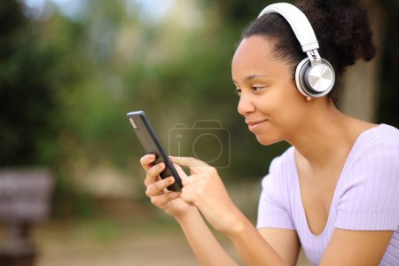 Téléchargez les photos : Femme noire portant un casque en utilisant le téléphone écoute audio dans un parc - en image libre de droit