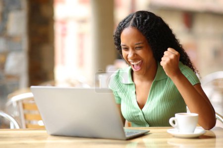 Téléchargez les photos : Femme noire excitée vérifiant les bonnes nouvelles sur un ordinateur portable dans une terrasse de bar - en image libre de droit