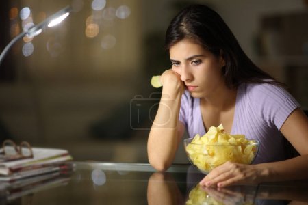 Foto de Triste mujer comiendo papas fritas quejándose en la noche en casa - Imagen libre de derechos