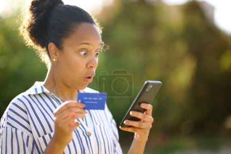 Photo for Surprised black buyer using phone and credit card to buy online in a park - Royalty Free Image