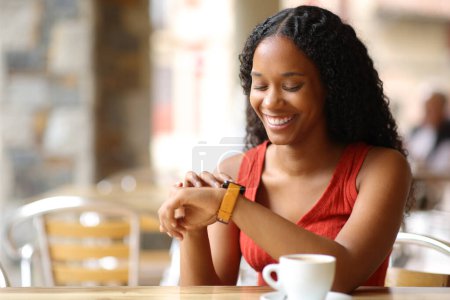 Foto de Mujer negra feliz comprobando smartwatch sentado en una terraza cafetería - Imagen libre de derechos