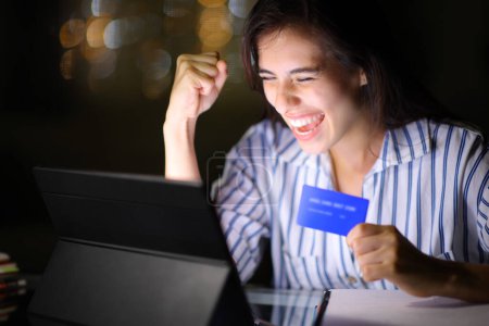 Photo for Excited woman buying online with tablet and credit card in the night at home - Royalty Free Image