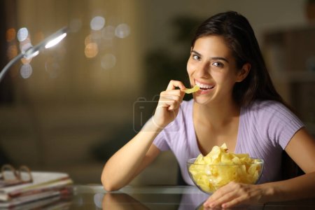 Foto de Mujer feliz en la noche comiendo papas fritas mirando a la cámara en casa - Imagen libre de derechos