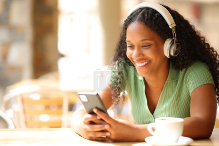 Foto de Mujer negra feliz escuchando música usando auriculares usando teléfono en una cafetería - Imagen libre de derechos