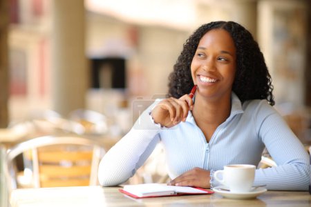 Foto de Mujer negra feliz con agenda pensando mirando al lado en una terraza del bar - Imagen libre de derechos