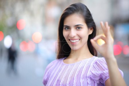 Téléchargez les photos : Femme heureuse gestuelle ok debout dans la rue vous regardant - en image libre de droit