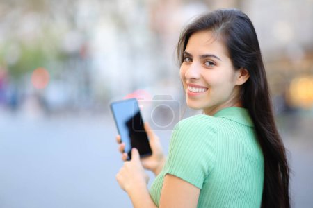 Téléchargez les photos : Femme heureuse tournoyant à l'aide d'un téléphone portable marchant dans la rue en regardant la caméra - en image libre de droit