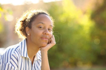Téléchargez les photos : Portrait d'une femme noire détendue assise dans un parc regardant la caméra - en image libre de droit