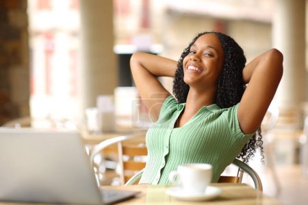 Téléchargez les photos : Femme noire heureuse détendue avec ordinateur portable reposant assis dans un bar terrasse vous regardant - en image libre de droit