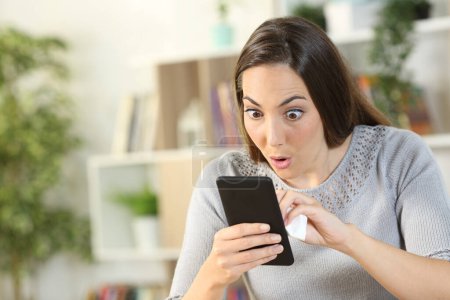 Téléchargez les photos : Femme surprise nettoyage téléphone intelligent avec les yeux et la bouche ouvert assis à la maison dans le salon - en image libre de droit