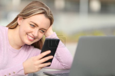 Téléchargez les photos : Happy teen vérifier le contenu du téléphone intelligent à côté d'un ordinateur portable dans la rue - en image libre de droit
