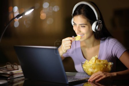 Téléchargez les photos : Femme heureuse dans la nuit portant un casque mangeant des croustilles de pomme de terre regarder les médias sur un ordinateur portable à la maison - en image libre de droit