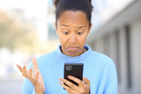 Téléchargez les photos : Portrait avant d'une femme noire perplexe vérifiant son téléphone portable dans la rue - en image libre de droit