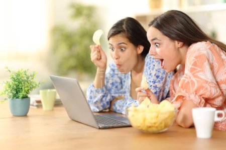 Photo for Surprised women watching media on laptop eating potato chips at home - Royalty Free Image
