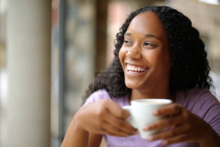 Téléchargez les photos : Heureuse femme noire buvant du café et riant en regardant côté dans une terrasse du restaurant - en image libre de droit