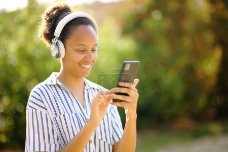 Téléchargez les photos : Heureuse femme noire écoutant audio avec casque et téléphone portable dans un parc - en image libre de droit