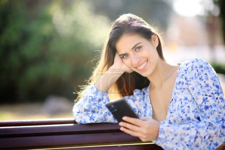 Téléchargez les photos : Femme heureuse vous regarde tenant le téléphone assis dans un parc - en image libre de droit