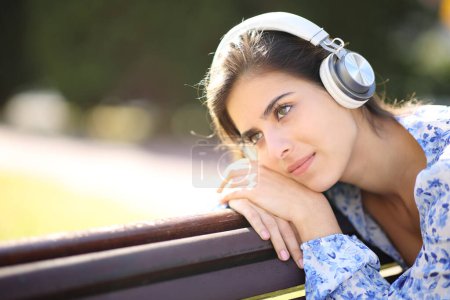 Foto de Mujer pensativa escuchando audio con auriculares en un parque - Imagen libre de derechos