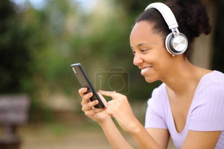 Téléchargez les photos : Heureuse femme noire portant un casque écoutant du contenu audio en utilisant le téléphone dans un parc - en image libre de droit