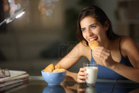Photo for Happy woman eating croissants and milk sitting in the night at home - Royalty Free Image