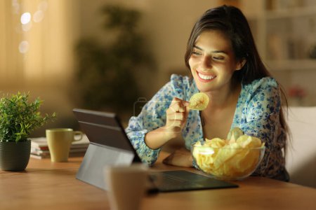 Téléchargez les photos : Femme heureuse dans la nuit regarder du contenu en ligne manger des croustilles à la maison - en image libre de droit