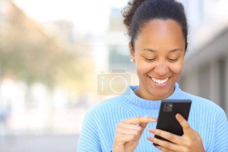 Téléchargez les photos : Portrait avant d'une femme noire heureuse utilisant un téléphone portable dans la rue avec espace de copie - en image libre de droit
