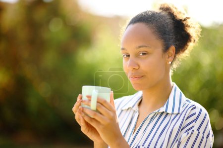 Photo for Serious black woman holding coffee cup looks at you in a park - Royalty Free Image