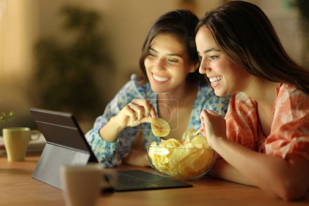 Foto de Dos amigos felices viendo películas en tabletas comiendo papas fritas en la noche en casa - Imagen libre de derechos