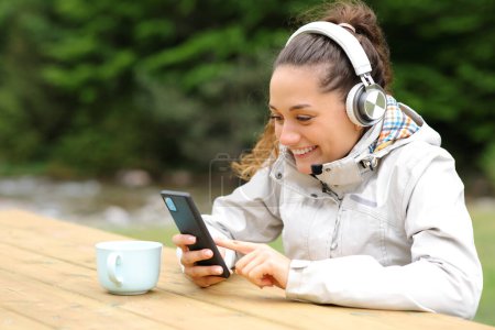 Photo for Happy hiker listening to music with headphones and smart phone in a picnic table - Royalty Free Image